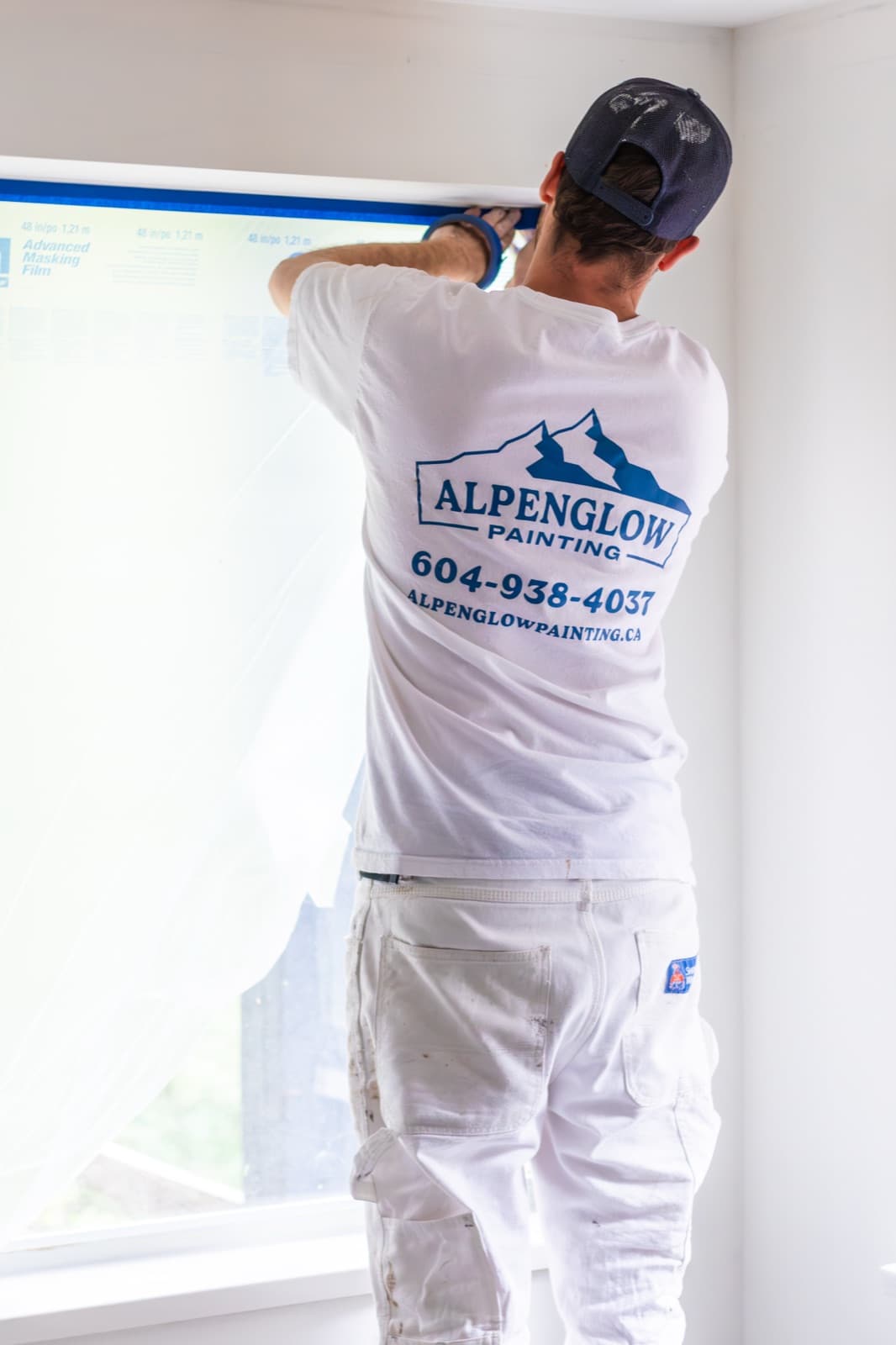 Noise-reduced version of a painter in an Alpenglow Painting branded white tee and navy cap applying blue tape and advanced masking film over a window