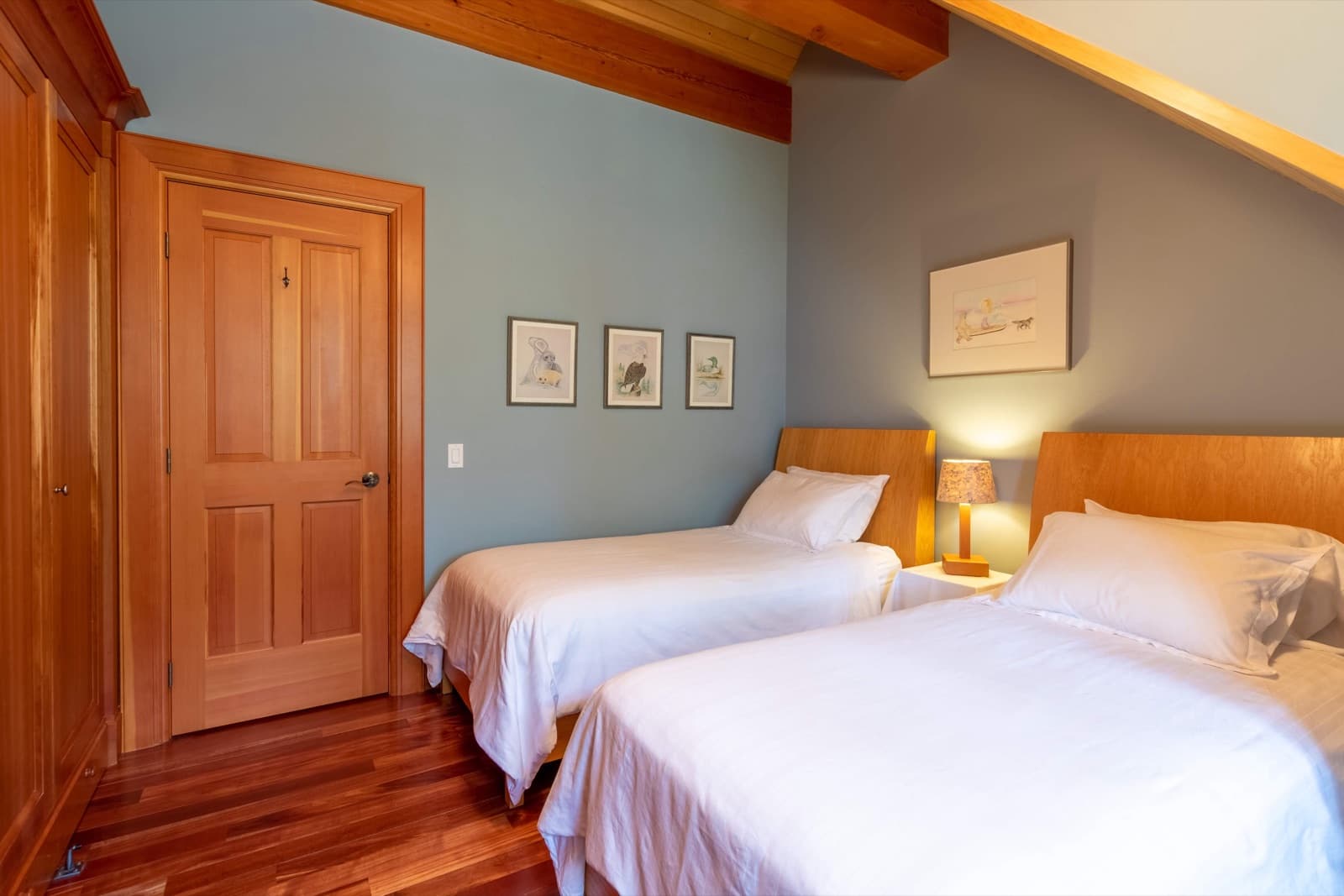 Attic guest bedroom with blue-grey painted walls, exposed fir beams, two twin beds with white linens and framed wildlife art prints
