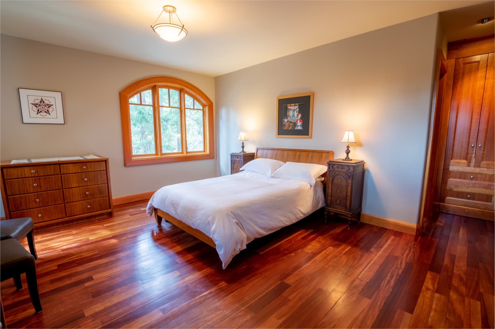 Master bedroom with soft grey painted walls, arched fir-trimmed window, glossy red-toned hardwood floor, white-linen bed and antique nightstands