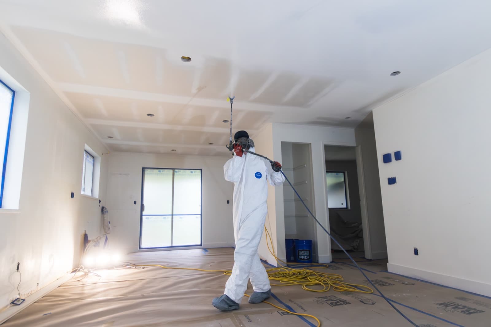 Painter in white coveralls walking while spraying a bright interior ceiling with an extension-pole airless sprayer, blue Sherwin-Williams PRO+ bucket