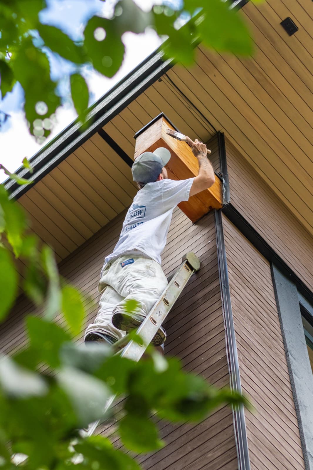 Painter on a ladder brush-applying clear stain to a fir timber box beam under the pale soffit of a modern brown-slat-sided home