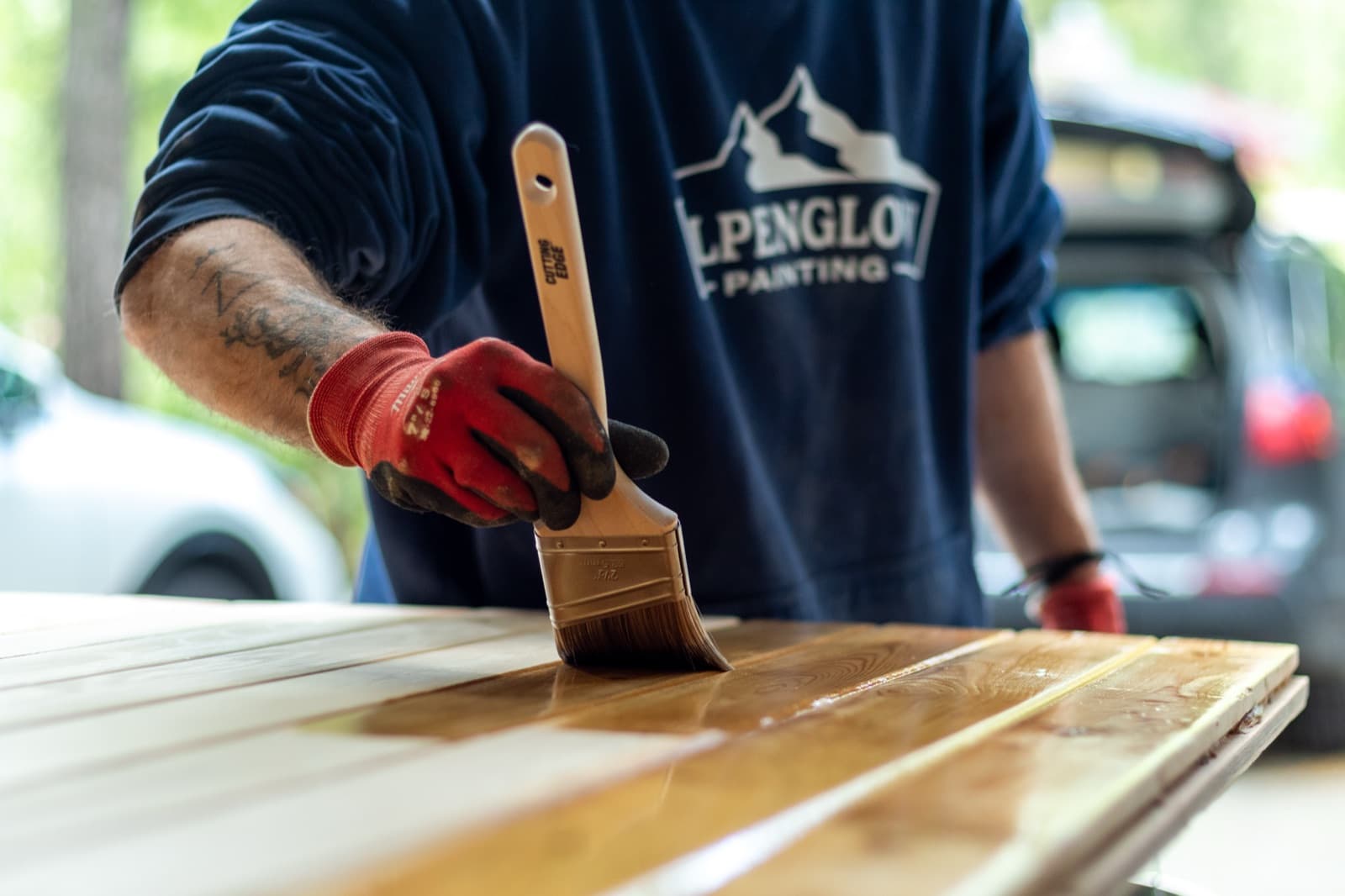 Close-up of an Alpenglow Painting crew member in branded navy sweatshirt and red gloves brushing golden stain onto cedar boards