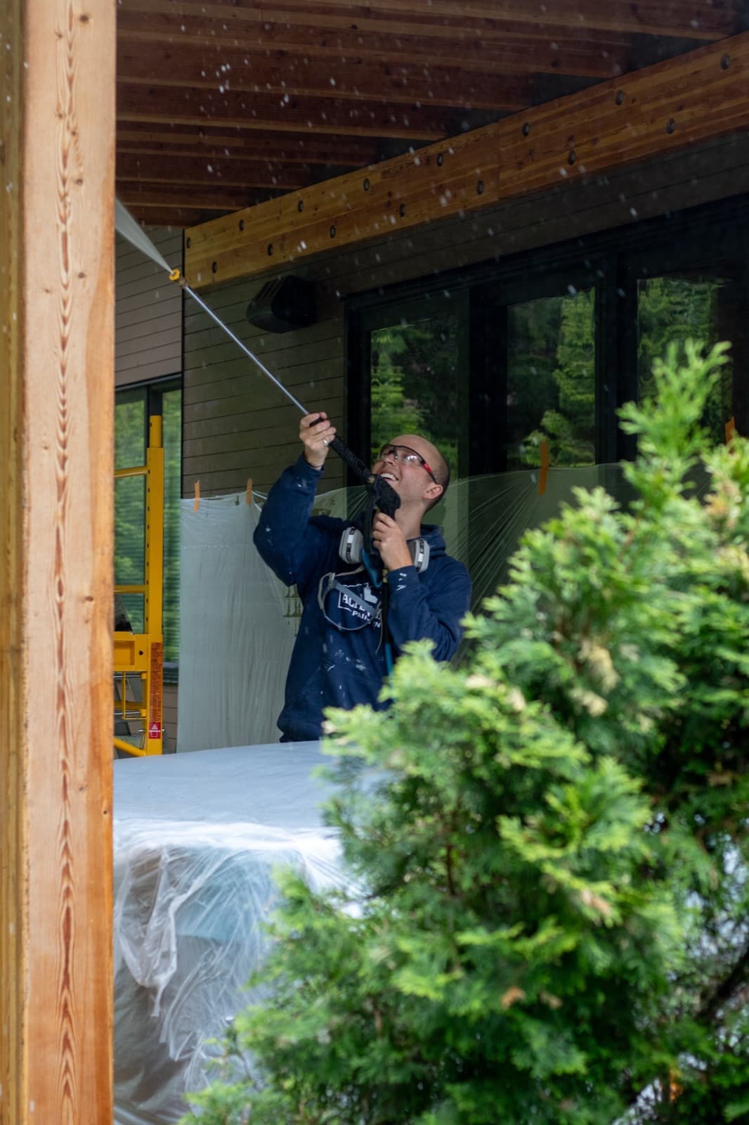 Smiling painter in a navy Alpenglow hoodie with respirator pressure-washes exposed timber soffit beams on a modern dark-sided chalet porch