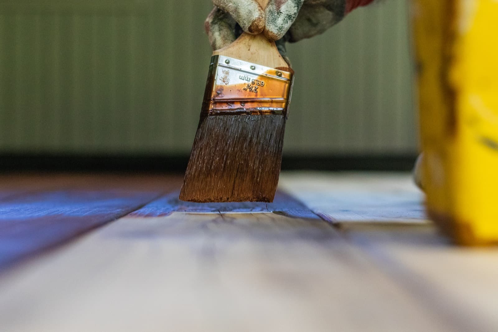 Low-angle macro of a 2.5-inch brush with bristles fully loaded in dark red-brown stain touching a cedar deck board