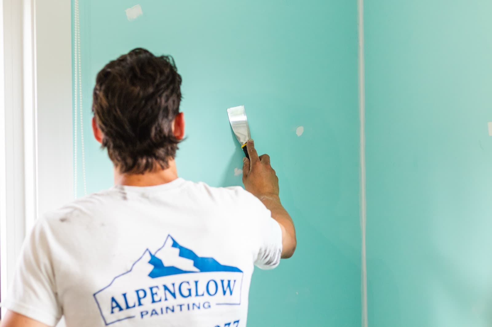Painter in an Alpenglow Painting shirt using a stainless putty knife to patch and prep a turquoise-aqua interior wall with white trim